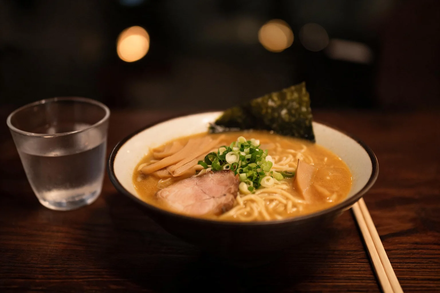 Bowl of ramen on a dark wooden table, glass of water and chopsticks, dim light (illustration)